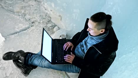 Man-is-sit-on-the-internet-in-laptop-in-an-ice-cave.-Around-the-mysterious-beautiful-ice-grotto.-User-communicates-in-social-networks.-He-crazy-strange-hipster-in-black-coat-and-fashionable-hairdo.