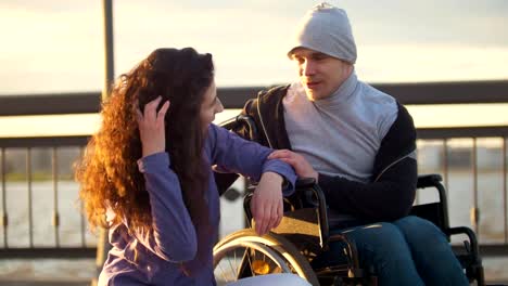 Happy-couple---disabled-man-in-wheelchair-talking-with-attractive-young-woman-enjoing-the-sunset