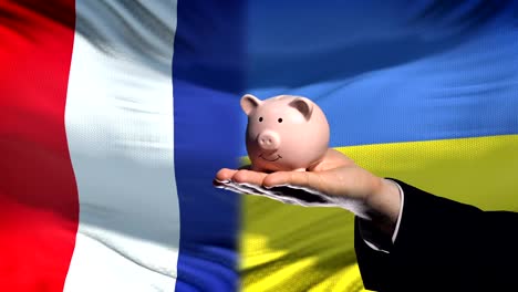 France-investment-in-Ukraine,-hand-putting-money-in-piggybank-on-flag-background