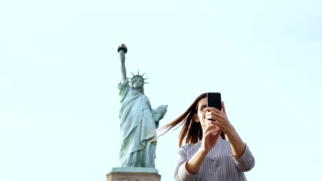 Schöne-glückliche-kaukasischen-touristischen-Frau-nimmt-ein-Selfie-Foto-mit-Smartphone-an-der-Freiheitsstatue-in-New-York-City