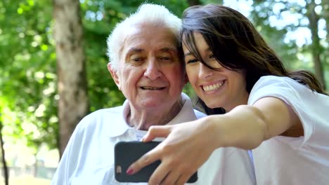 Una-niñera,-enfermera,-cuidar-de-los-ancianos,-una-chica-(mujer)-y-el-abuelo-se-sientan-en-un-libro-de-bolsillo-y-hacer-selfie,-en-el-parque.