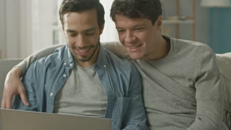 Adorable-Male-Gay-Couple-Spend-Time-at-Home.-They-Sit-on-a-Sofa-and-Use-the-Laptop.-They-Watch-Funny-Online-Videos.-Partner-Puts-His-Hand-Around-His-Lover.-Room-Has-Modern-Interior.