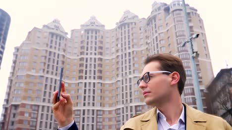 Closeup-portrait-of-attractive-caucasian-man-taking-video-of-the-city-on-the-phone-on-the-background-or-urban-high-buildings