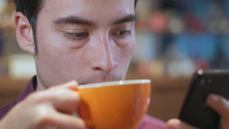 Un-atractivo-guapo-guapo-hispano-América-Latina-joven-es-utilizando-su-teléfono-celular-y-tomar-una-taza-de-café-en-una-cafetería,-Close-up-extremo,