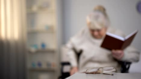 Old-woman-sitting-in-wheelchair-reading-book,-searching-eyeglasses,-bad-vision