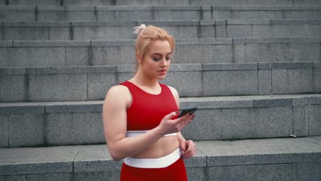 Plus-size-blond-mixed-racee-smiling-woman-wearing-red-sportswear-using-smartphone-outdoors