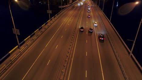 Volando-sobre-el-tráfico-de-coches-por-la-noche-en-un-puente-sobre-un-río.-Plano-aéreo,-UHD