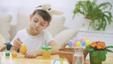 Intersted-and-concentrated-boy-has-finished-colourizing-an-Easter-egg-in-an-yellow-colour,-sitting-at-the-table-and-tasting-an-orange-lollipop.