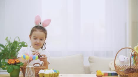 Litle-playful-girl,-wearing-bunny-ears-on-her-head-is-choosing-an-Easter-egg.-Suddenly,-somebody-noticed-her.-She-got-frightened-and-hid-down.