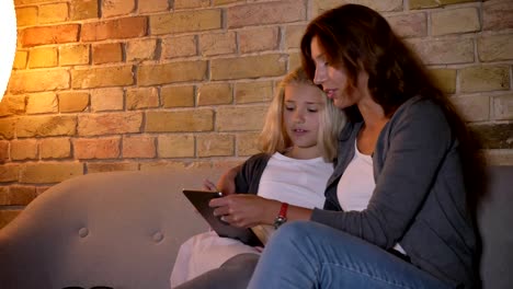 Closeup-Shooting-der-jungen-kaukasischen-Mutter-und-ihres-kleinen-hübschen-blonden-Mädchens,-das-die-Tablette-zusammen-benutzt,-während-sie-auf-der-Couch-sitzt-und-umarmt