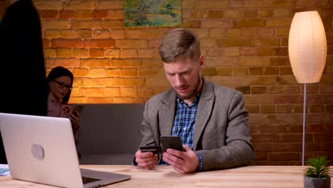 Closeup-shoot-of-adult-businessman-shopping-online-on-the-phone-in-front-of-the-laptop-indoors-in-the-office.-Female-employee-using-the-cellphone-on-the-background