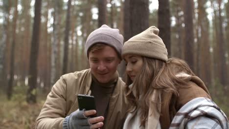 Cheerful-Couple-Using-Smartphone-and-Chatting-in-Forest-on-Autumn-Day