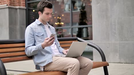 Young-Man-sitzt-mit-Smartphone-und-Laptop-im-Freien