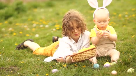 Two-brothers,-a-teenager-and-a-baby-are-resting-in-a-park-on-a-glade.-Easter-concept,-bunny-ears,-easter-basket-and-eggs.-A-dog-runs-past-them