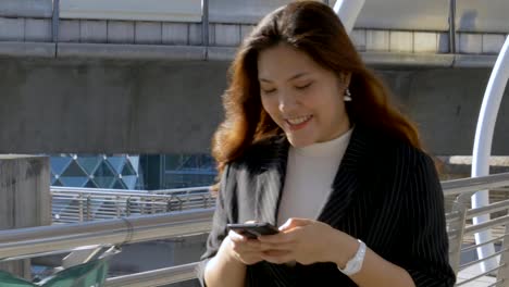 Lächelnde-junge-asiatische-Frau,-die-mit-Freund-auf-dem-Smartphone-am-städtischen-Stadthintergrund-unterwegs-ist,-sucht-nach-sozialen-Medien.