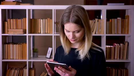 Nahaufnahme-von-jungen-attraktiven-Studentin-mit-dem-Tablet-Blick-auf-die-Kamera-in-innenräumen-in-der-Universitätsbibliothek