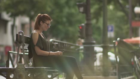 Attractive-redhead-woman-with-glasses,-freckles,-piercings-and-red-hair-writing-a-text-message-on-her-smartphone-sitting-on-street-bench,-during-sunny-summer-in-Paris.-Slow-motion.-Medium-Shot.