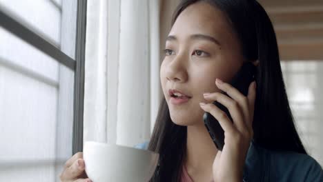 Close-up-attractive-portrait-smiling-young-asian-woman-drinking-coffee-and-talking-on-Phone-with-friends-standing-beside-window-at-home-office.