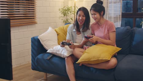 Lesbian-lgbt-women-couple-play-games-at-home,-Asian-female-using-joystick-having-funny-happy-moment-together-on-sofa-in-living-room-in-night.-Young-lover-football-fan,-celebrate-holiday-concept.