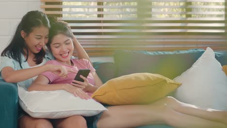 Young-Asian-Lesbian-women-couple-using-mobile-phone-check-social-media-on-internet-together-while-lying-sofa-in-living-room.