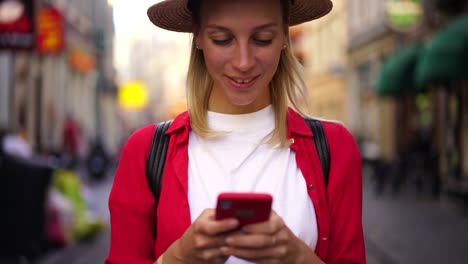 Mujer-caucásica-positiva-en-sombrero-leer-mensaje-en-el-teléfono-móvil-usando-la-aplicación-en-línea-en-la-calle-de-la-ciudad