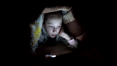 A-little-girl-playing-on-a-tablet-at-night-on-a-bed-under-a-blanket.-Concept-video.-Close-up.-Raw-video.-4K.