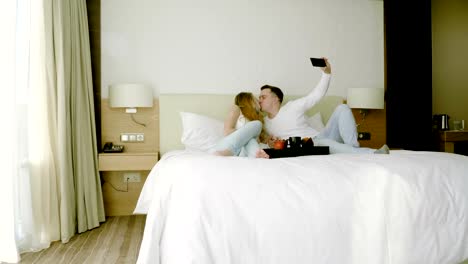 Happy-couple-looking-at-cell-phone,-kissing,-taking-a-selfie-in-bedroom.-4K