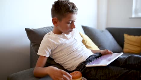 Young-boy-is-playing-games-on-tablet-and-eating-doughnut