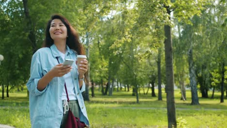 Beautiful-Asian-woman-walking-in-park-with-smartphone-and-to-go-coffee-smiling