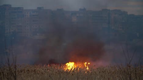 Vista-impactante-de-bloques-de-apartamentos-multistoried-en-la-orilla-del-río-Dnipro-y-el-fuego-resplandeciente-a-través-del-río-en-el-pantano-de-caña-en-primavera