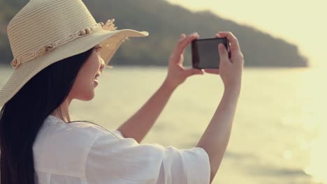 Schöne-asiatische-Frau-hält-Smartphone,-ein-Foto-am-Strand-schönen-Sonnenuntergang.-Lifestyle-Reise-Urlaub-Urlaub-Sommerkonzept.