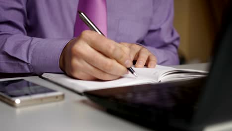 Businessman-in-a-purple-shirt-and-tie-makes-notes-in-a-notebook.-Man-writes-a-pen-in-daily.-A-person-makes-notes-in-organizer.-Laptop-and-smartphone-on-office-desk-in-office.-Closeup.