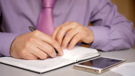 Businessman-in-a-purple-shirt-and-tie-makes-notes-in-a-notebook.-Man-writes-a-pen-in-daily.-A-person-makes-notes-in-organizer.-Laptop-and-smartphone-on-office-desk-in-office.-Closeup.