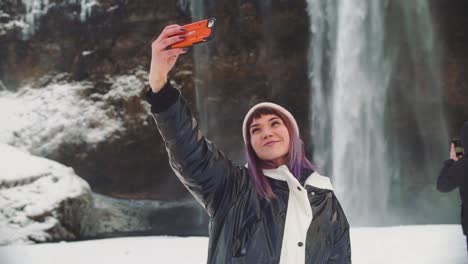 beautiful-young-woman-near-a-waterfall-in-iceland,-takes-pictures-on-the-phone,-rejoices-and-takes-a-selfie