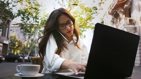 Business-female-in-glasses,-white-shirt.-Sitting-at-table-with-cup-of-coffee-and-laptop-in-roadside-cafe.-Talking-on-mobile-phone,-typing.-Slow-motion