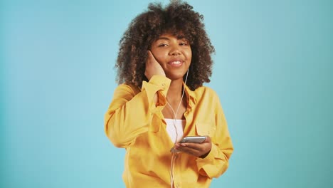 Mujer-afroamericana-escuchando-música-a-través-de-auriculares-conectados-a-teléfonos-inteligentes,-bailando-y-sonriendo-posando-sobre-fondo-azul