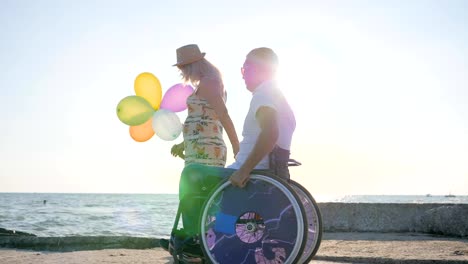 happy-disabled-Daddy-walking-with-together-with-family-beside-sea-in-backlight,-kid-with-colorful-air-balloons-walks