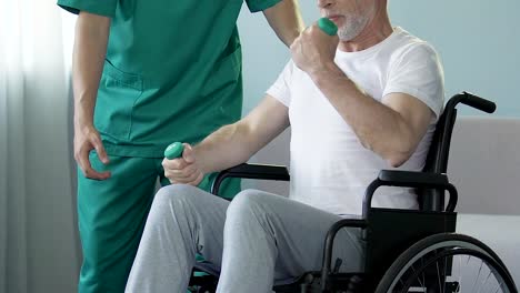 Senior-man-in-wheelchair-flexing-arms-with-dumbbells,-assisted-by-nurse,-rehab