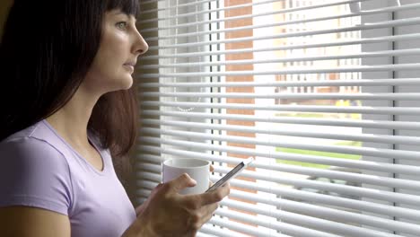 Joven-bebe-pie-café-con-smartphone-por-ventana-en-la-oficina.-Niña-mira-por-la-ventana-y-utiliza-un-smartphone.-Mensaje-de-texto,-navegar-por-Internet,-aplicación,-red-social.