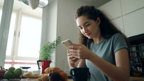 young-beautiful-curly-pretty-caucasian-woman-sitting-at-table-in-nice-kitchen-using-smartphone,-she-is-texting-someone-and-smiling,-calm-and-happy