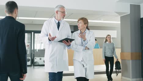 In-the-Hospital,-Busy-Doctors-Talk,-Using-Tablet-Computer-While-Walking-Through-the-Building.-In-the-Background-Patient-Talks-with-Receptionist.-New-Modern-Fully-Functional-Medical-Facility.