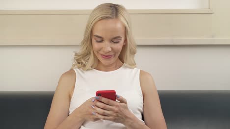 A-close-up-of-a-lovely-blonde-sitting-on-the-sofa,-looking-at-the-phone,-smiling.-The-camera-moves-from-the-left-to-the-right.