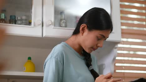 Mujer-asiática-joven-usando-la-tableta-y-comiendo-pan.-tecnología,-red-social,-concepto-de-comunicación-en-la-cocina-en-casa.