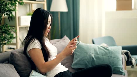 Attractive-Asian-lady-is-using-smartphone-to-make-online-payment-with-bank-card-sitting-on-sofa-at-home-and-shopping-in-the-internet.-Banking-and-youth-concept.