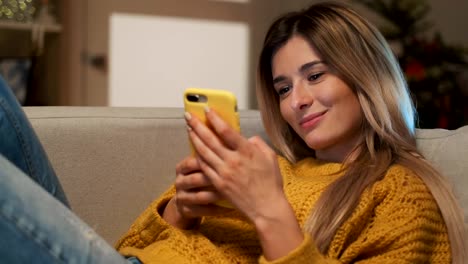 Attractive-blonde-student-girl-texting-on-her-phone.-Woman-using-smartphone-to-communicate-with-friends-in-social-network-typing-messages-and-looking-through-internet-sitting-on-comfort-sofa-at-home