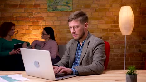 Closeup-shoot-of-adult-businessman-typing-on-the-laptop-looking-at-camera-and-smiling-indoors-in-the-office.-Female-employee-discussing-data-with-a-colleague-on-the-background