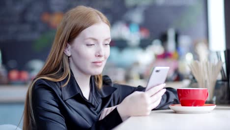 Zoom-in-of-cheerful-young-woman-with-red-hair-sitting-at-counter-in-cafe-with-cup-of-coffee-nearby-and-browsing-social-networks-with-smile