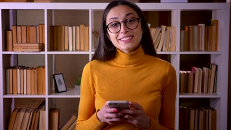 Retrato-de-hermosa-maestra-morena-en-gafas-mirando-en-el-teléfono-inteligente-de-pie-con-atención-en-la-biblioteca.