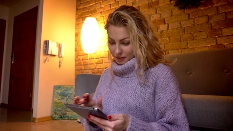 Closeup-shoot-of-adult-caucasian-blonde-female-using-the-tablet-while-sitting-on-the-floor-and-leaning-on-the-couch-indoors-in-a-cozy-apartment