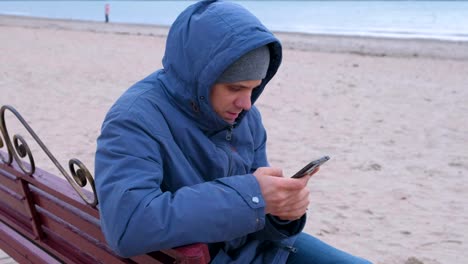 Man-blogger-in-a-blue-down-jacket-sitting-on-a-bench-on-the-sand-beach-and-writing-a-post-in-social-media-on-mobile-phone.